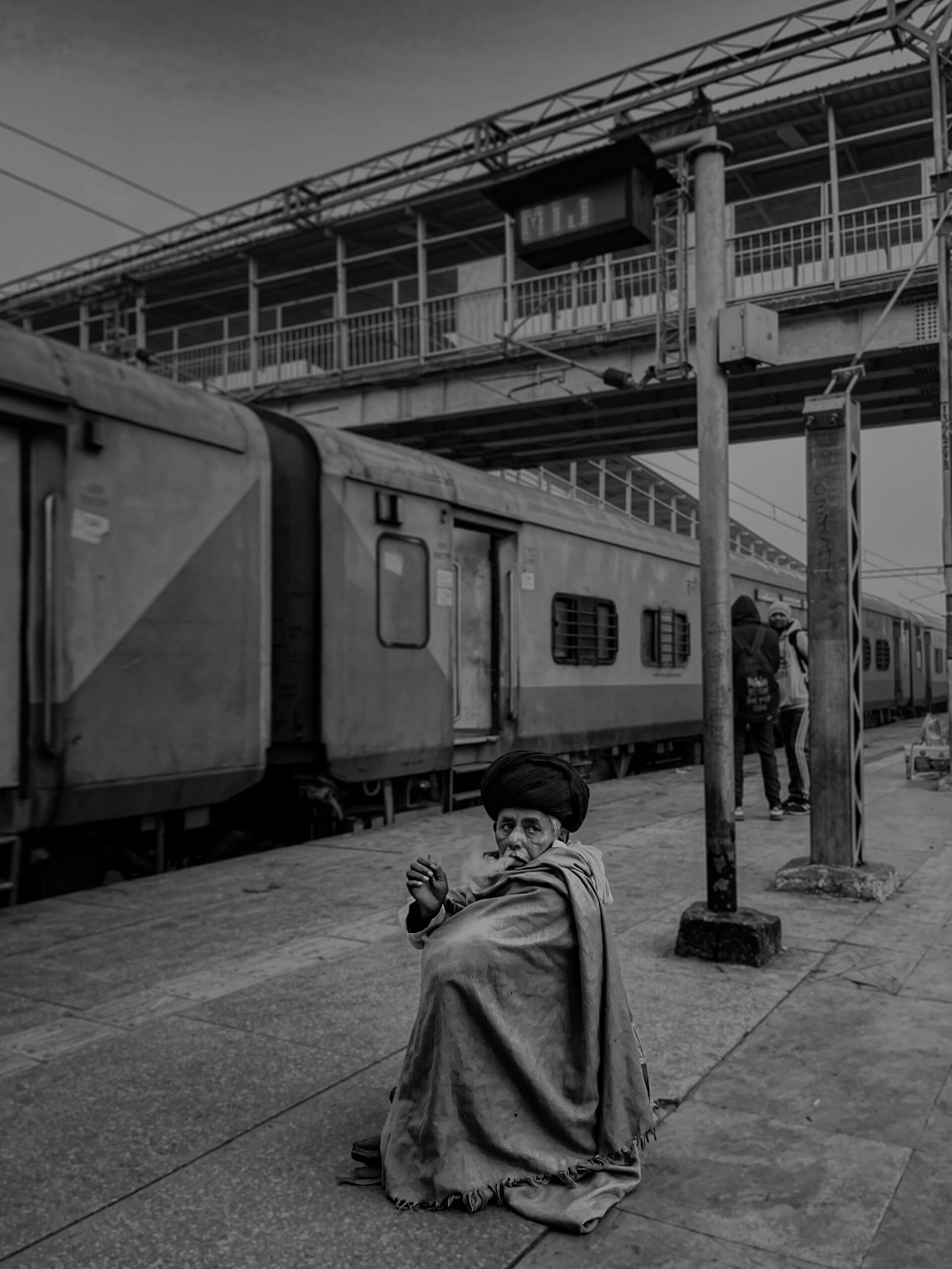 Black and white image of a person sitting on the platform at Mathura railway station, India.