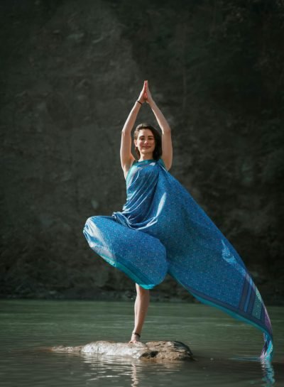 A woman in a blue dress performs a yoga tree pose on a rock in a river at Rishikesh, India.